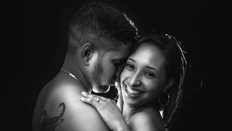 Pareja abrazada en blanco y negro sobre fondo oscuro; ella sonríe a cámara con aros y anillo, él lleva un tatuaje